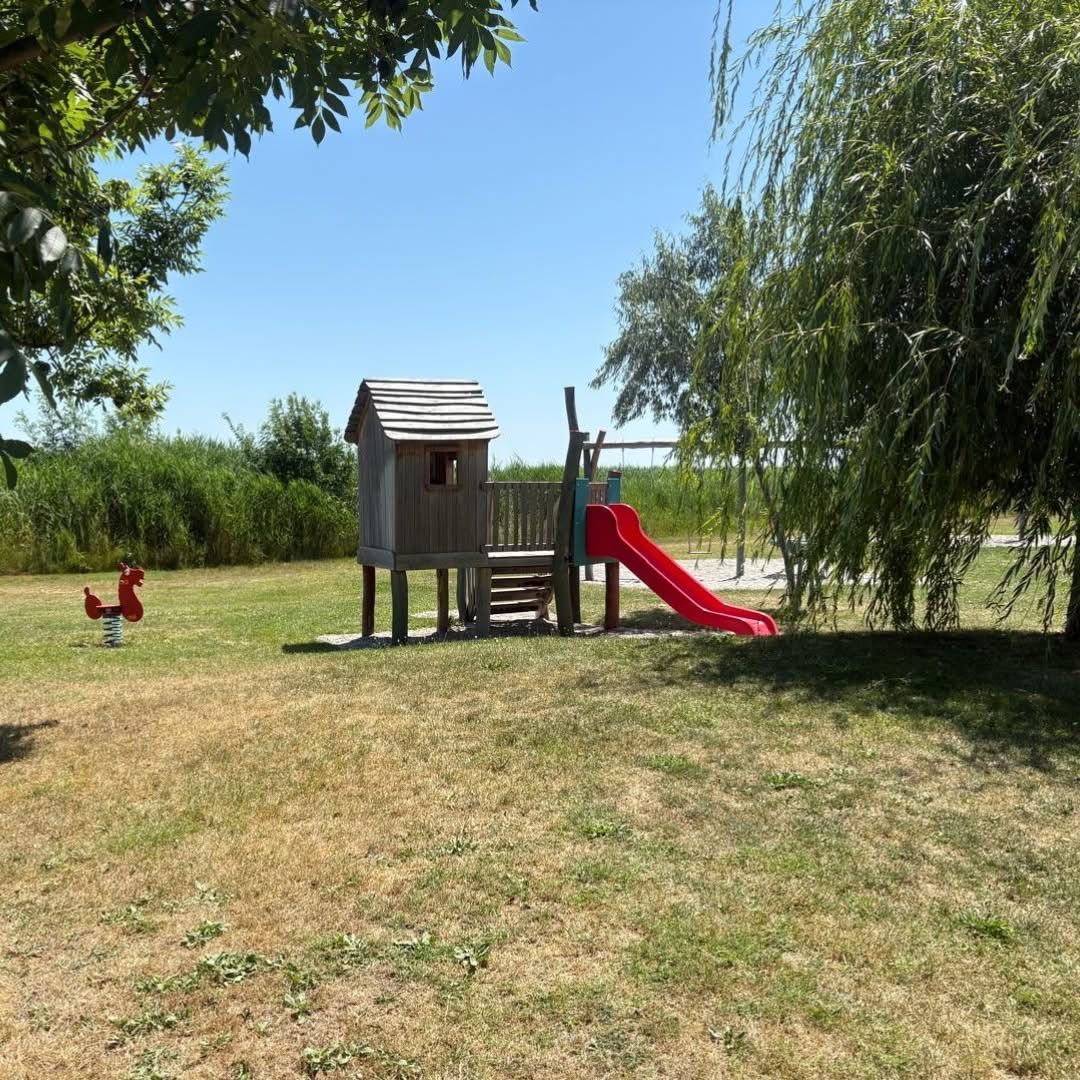 Ein Spielplatz mit einem Holzhaus, einer roten Rutsche und einer Schaukel befindet sich in einem Grasbereich unter einem Baum.