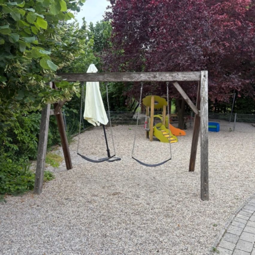 Ein Spielplatz mit einem hölzernen Schaukelgerüst und einer Rutsche, umgeben von Bäumen und Kies.