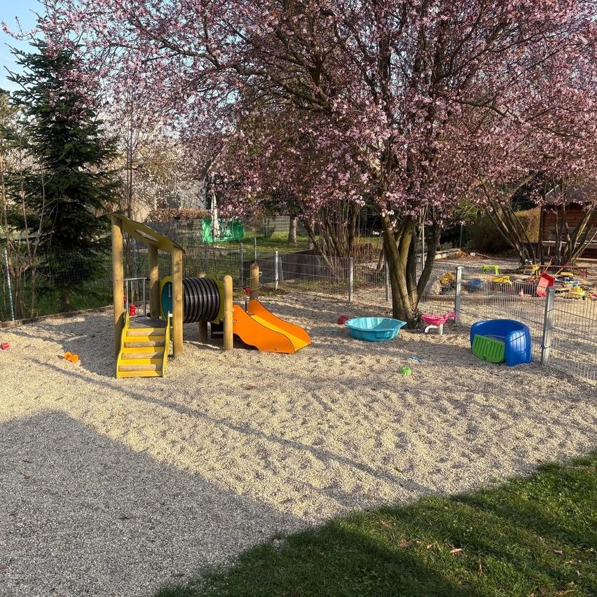 Ein Kinderspielplatz mit einer Rutsche, Stufen und verschiedenen Spielzeugen in einem sandigen Bereich, umgeben von Bäumen, Gras und einem Zaun.