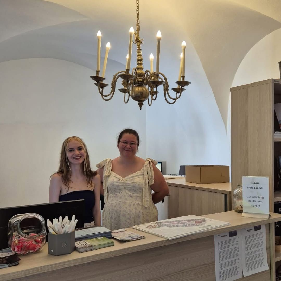 Zwei Frauen stehen hinter einer Rezeption. Eine Kronleuchter hängt an der Decke. Papiere und Bücher liegen auf dem Schreibtisch, zusammen mit einer Schachtel Süßigkeiten. Ein Schild zeigt 'Spenden' und 'Danke!' an.