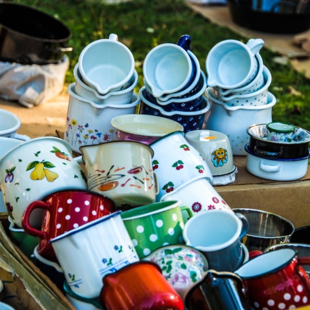 Eine Sammlung von Tassen und Bechern auf einem Holztisch, einige mit Blumendesigns, andere mit Punkten. Verschiedene Farben sind vorhanden, einschließlich Rot, Blau, Grün und Weiß. Ein Korb mit Tassen befindet sich auf der linken Seite.