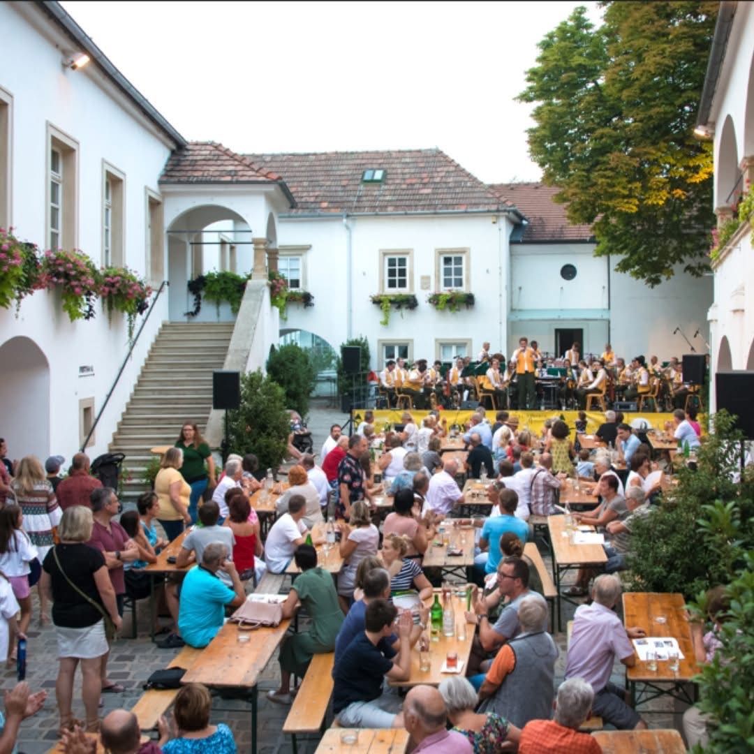 Eine Gruppe von Menschen sitzt im Freien an Tischen rund um einen Innenhof. Eine Band spielt auf einer Bühne mit gelbem Hintergrund vor einem weißen Gebäude.