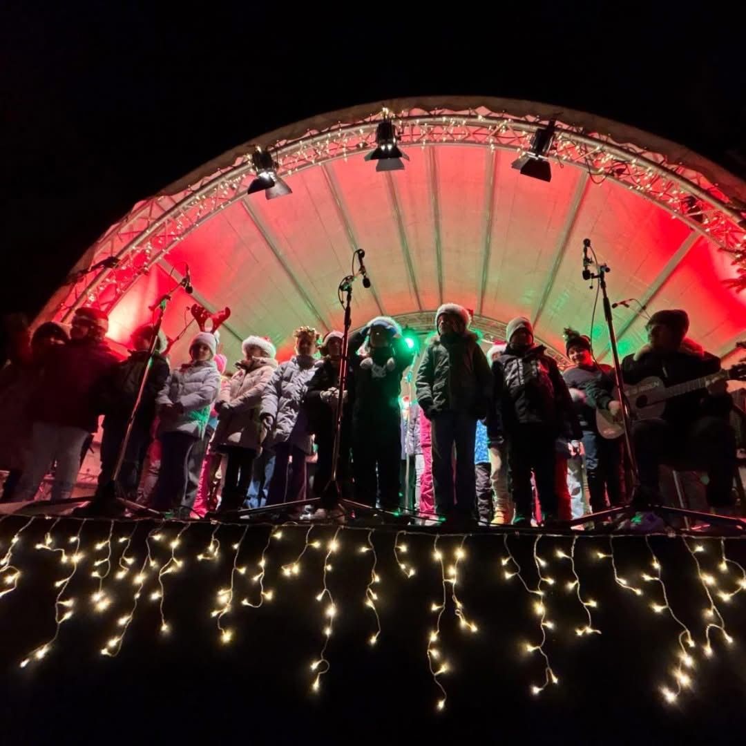 Eine Gruppe von Menschen steht mit Gitarren und Mikrofonen auf der Bühne und performt unter einem rot-grünen Bogen bei Nacht. Sie sind für das kalte Wetter warm gekleidet.