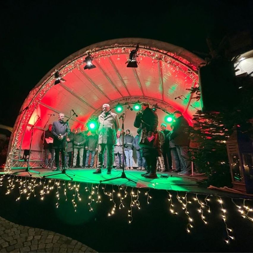 Eine nächtliche Aufführung auf einer Bühne mit einem festlichen roten und grünen Baldachin. Mehrere Personen stehen auf der Bühne, darunter eine mit einem Mikrofon. Die Bühne ist mit Weihnachtslichtern beleuchtet.