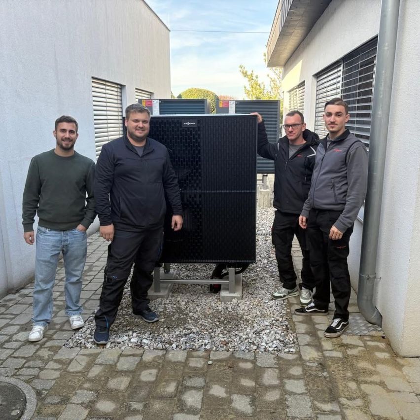 Vier Männer stehen vor einem Gebäude mit einem großen schwarzen Solarpanel und lächeln für ein Foto.