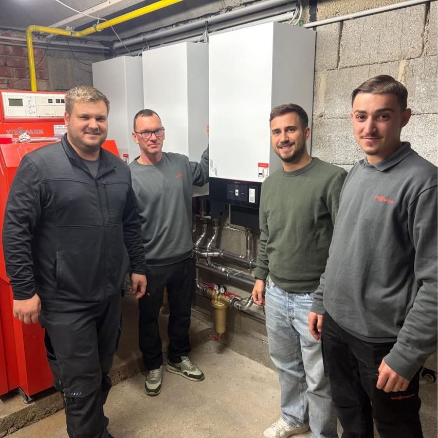 Vier Männer stehen vor einer Wand mit einer rot-weißen Maschine. Sie tragen graue, grüne und schwarze Jacken.