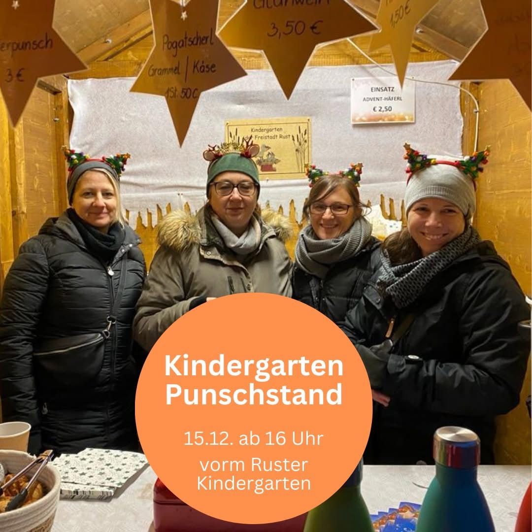 Fünf Erwachsene mit Weihnachtskopfbändern posieren für ein Foto. Dahinter zeigt ein Holzstand verschiedene Lebensmittelpreise. Rechts weist ein Schild auf einen Advent-Haferl-Preis von 2,50 Euro hin. Unten markiert ein orangefarbener Kreis eine Kindergarten-Punschstand-Veranstaltung, geplant für den 15.12. um 16 Uhr.