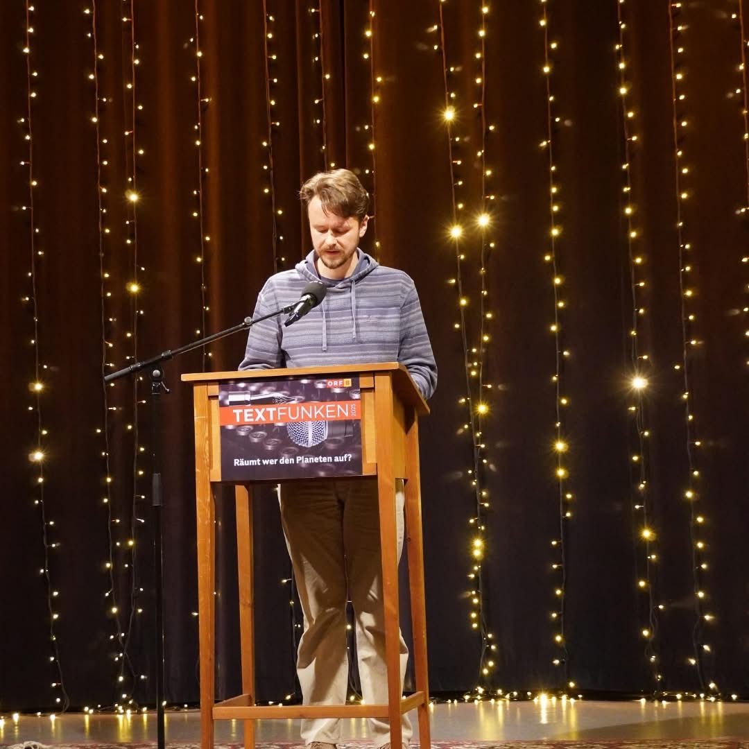 Ein Mann steht an einem Rednerpult mit einem Mikrofon und hält eine Rede oder Präsentation. Dahinter befindet sich ein Hintergrund mit Lichterketten.