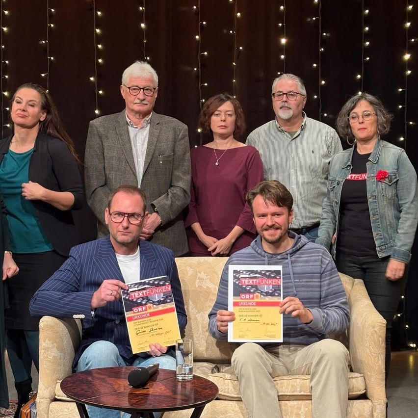 Eine Gruppe von Menschen steht und sitzt auf einer Couch in einem Studio und hält Urkunden. Sie alle lächeln und posieren für ein Foto. Ein Mikrofon steht auf dem Tisch vor ihnen.