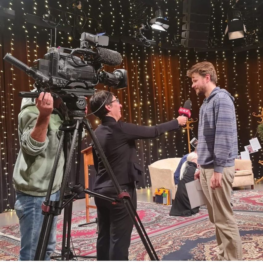 Ein Mann wird auf einer Bühne von einer Frau mit einem Mikrofon interviewt. Hinter ihnen steht eine Kamera auf einem Stativ. Eine andere Person hält die Kamera. Im Hintergrund befinden sich ein Sofa, ein Schreibtisch und Lichterketten.