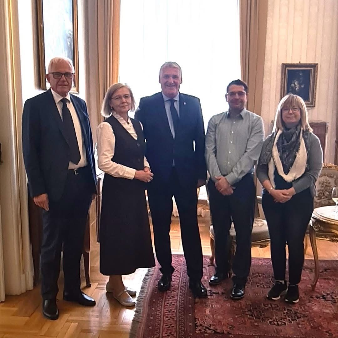 Fünf Personen stehen in einem Raum mit Holzboden und einer weißen Gardine hinter ihnen. Ein Mann in Anzug und Krawatte steht in der Mitte.
