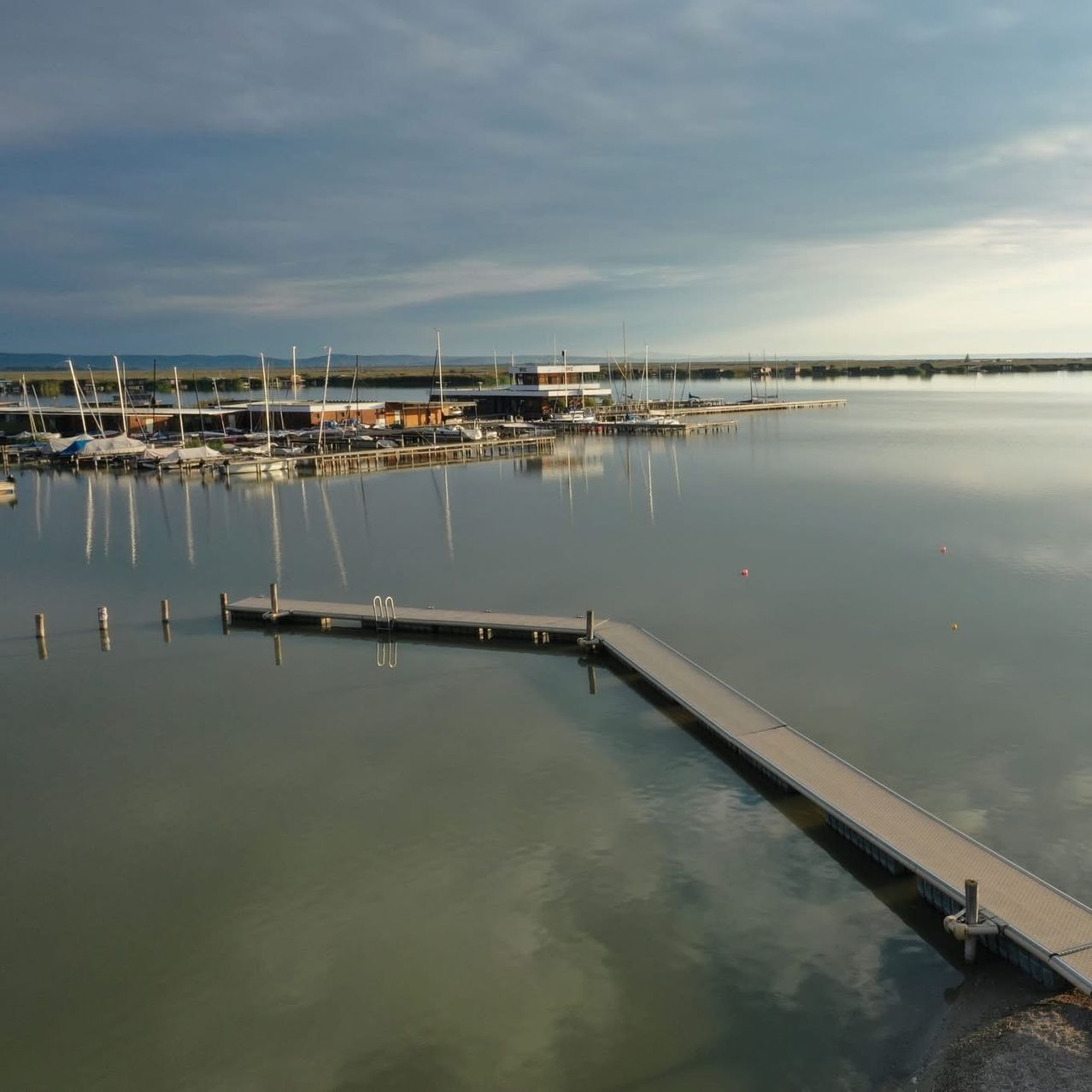 Eine Luftaufnahme eines Hafens mit angedockten Booten, einem Gebäude in der Ferne und einem langen Steg, der sich in ruhiges Wasser unter einem bewölkten Himmel erstreckt.
