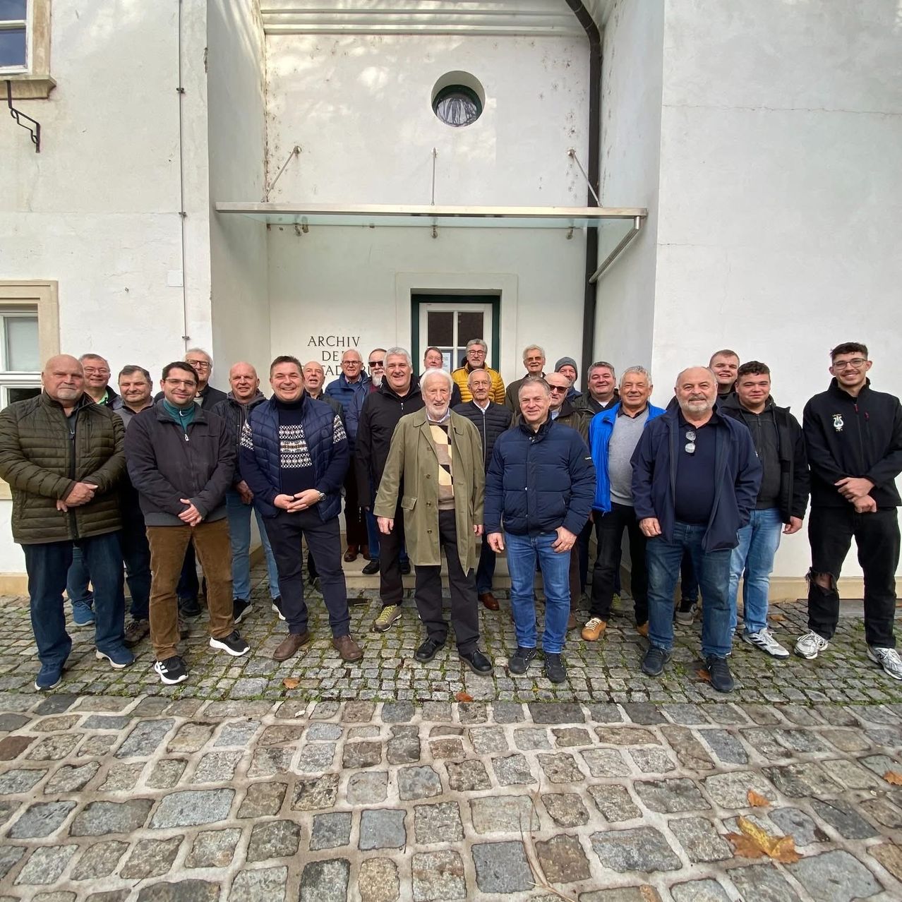 Eine Gruppe von Männern steht vor einem weißen Gebäude mit dem Schild 'ARCHIV DE'. Sie tragen Winterkleidung und scheinen für ein Foto zu posieren.