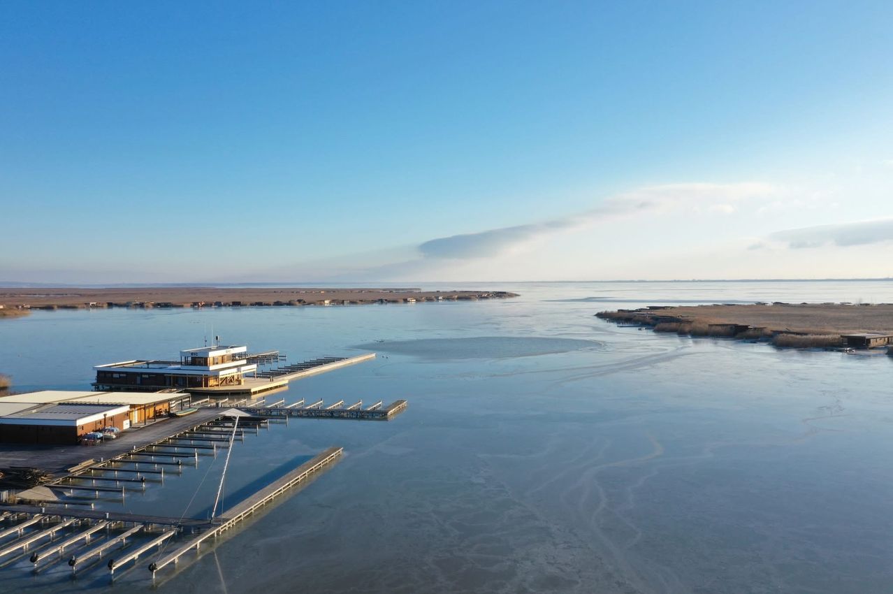 Eine Luftaufnahme eines ruhigen Meeres mit einem Pier und Gebäuden. Ein bewölkter Himmel mit einem blauen Farbton.