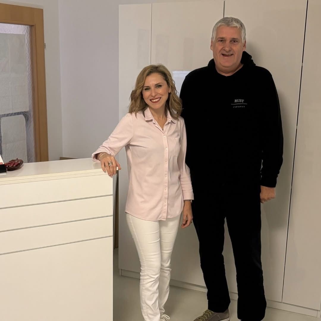 Ein Mann und eine Frau stehen lächelnd vor einem weißen Schrank. Die Frau trägt eine rosa Bluse und weiße Hose, der Mann ein schwarzes Hoodie. Dahinter ist eine weiße Wand mit einer Holztür.