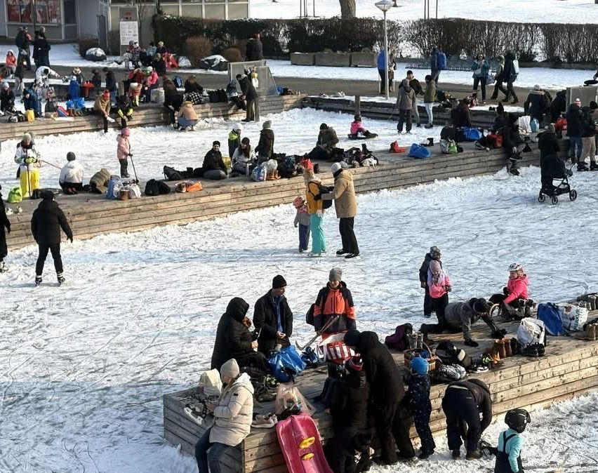 Eine Menschenmenge versammelt sich auf einer verschneiten Oberfläche. Sie sitzen und stehen auf Holzbänken. Einige tragen Mützen, Handschuhe und Stiefel. Mehrere Menschen tragen Taschen. Einer schiebt einen Kinderwagen. Ein Laternenpfosten ist in der Ferne.