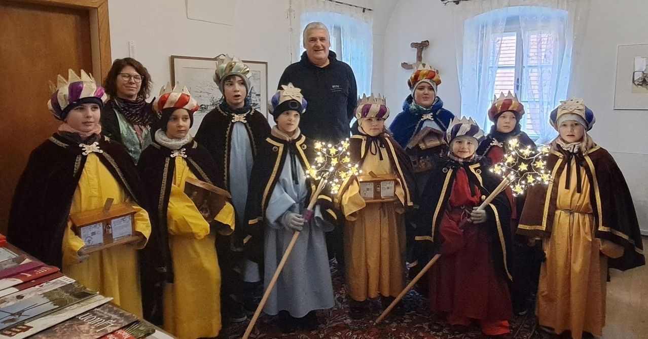 Eine Gruppe von Kindern, die als Könige gekleidet sind, steht zusammen mit einem Mann in einem schwarzen Jacken und hält einen beleuchteten Weihnachtsbaum. Hinter ihnen ist eine weiße Wand mit einem gerahmten Bild und einem Holzkreuz.