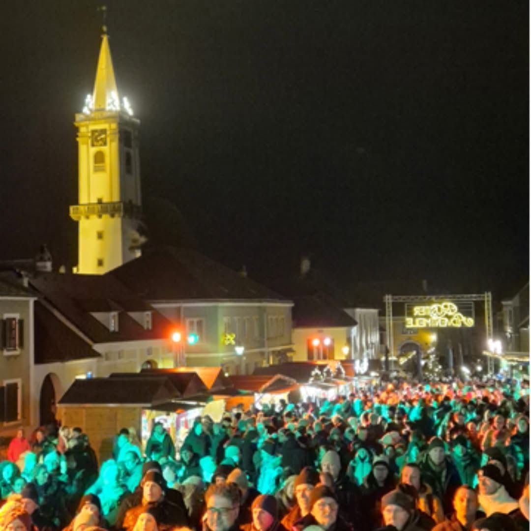 Bei Nacht versammelt sich eine große Menschenmenge auf einem Stadtplatz, der von festlichen Lichtern erleuchtet wird. Ein Turm mit einer Uhr ist im Hintergrund zu sehen.