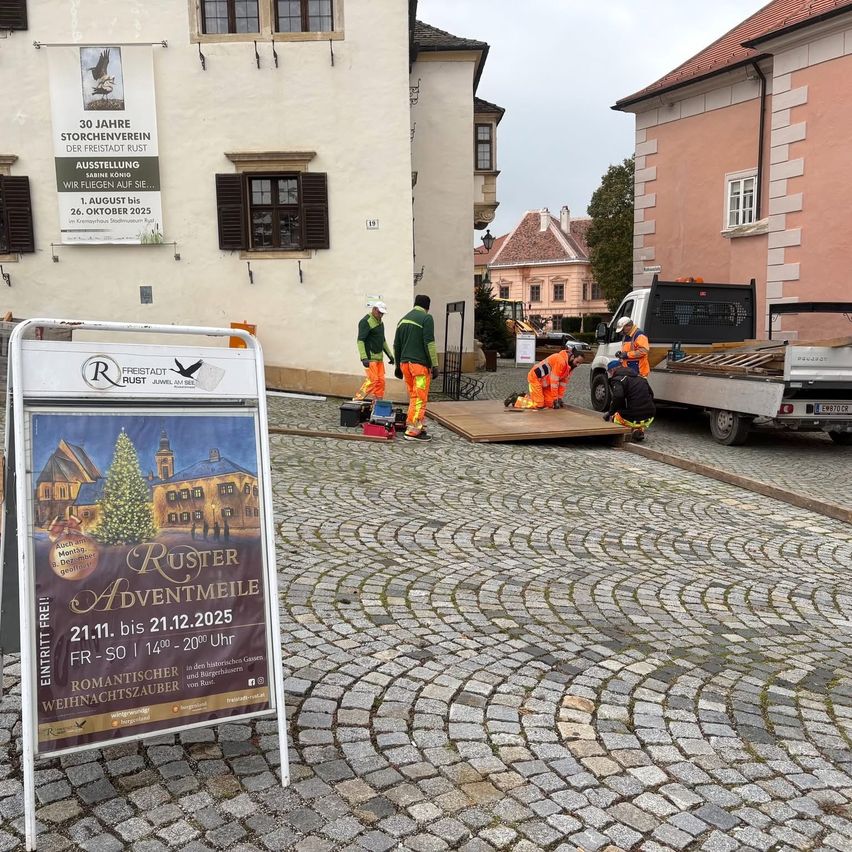 Ein Schild für Ruster Adventmeile steht auf einer Kopfsteinpflasterstraße. Arbeiter installieren eine Holzplattform. Dahinter ist ein Lastwagen geparkt. Auf beiden Seiten stehen Gebäude.