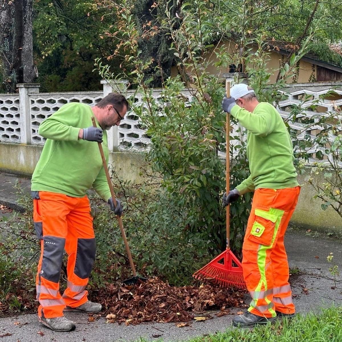 Zwei Arbeiter säubern die Blätter auf dem Bürgersteig. Einer benutzt eine Harke und der andere einen Besen. Beide tragen grüne und orange Uniformen.