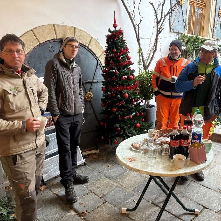 Vier Männer stehen in einem Innenhof mit einem Weihnachtsbaum. Sie tragen Winterjacken und Mützen. Der Tisch hat Tassen, Gläser und einen Korb. Ein Mann hält eine Tasse, und ein anderer hat eine Zigarette.