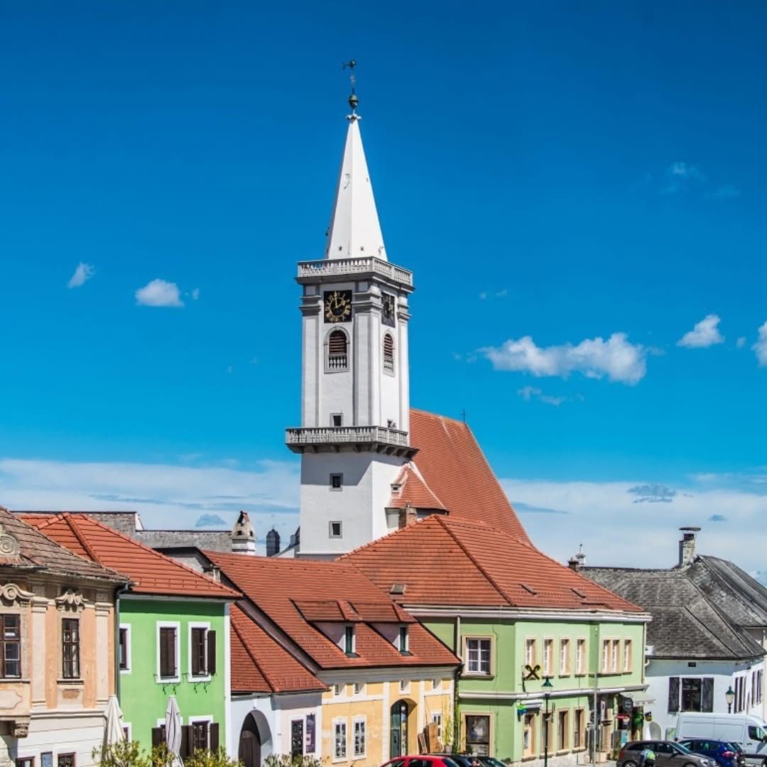 Ein Stadtplatz mit einer Kirche, die einen hohen Turm und eine Uhr hat, umgeben von farbenfrohen Gebäuden unter einem blauen Himmel mit Wolken.
