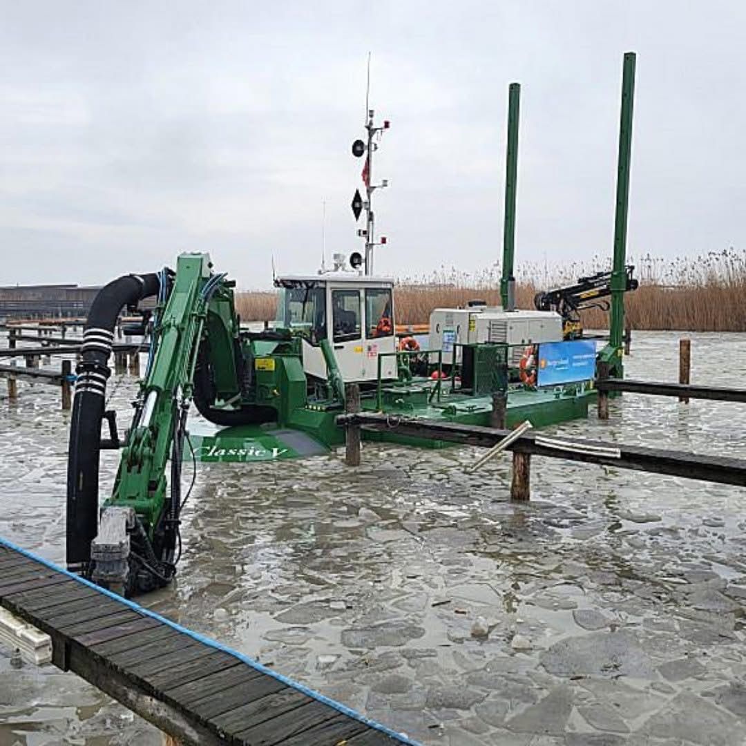 Ein grünes Wasserfahrzeug mit einem Kran ist in einem Gewässer verankert. Der Kran ist ausgestreckt und hat ein Schild.
