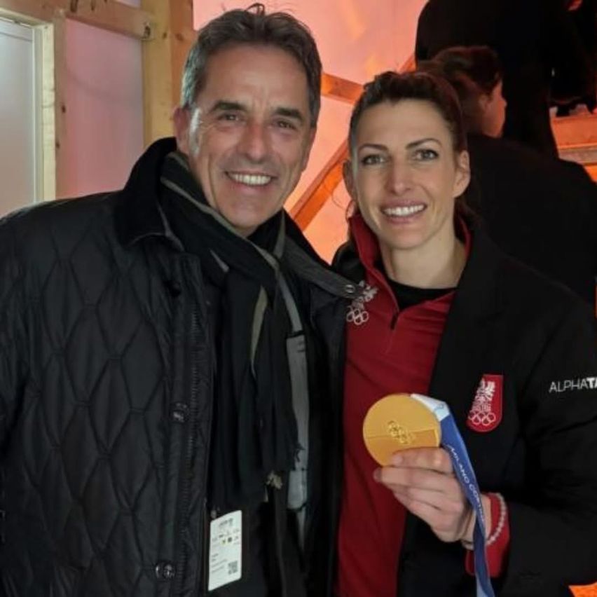 Ein Mann und eine Frau stehen lächelnd für ein Foto. Die Frau hält eine Goldmedaille. Sie tragen beide schwarze Jacken. Die Frau hat ein rotes T-Shirt darunter.
