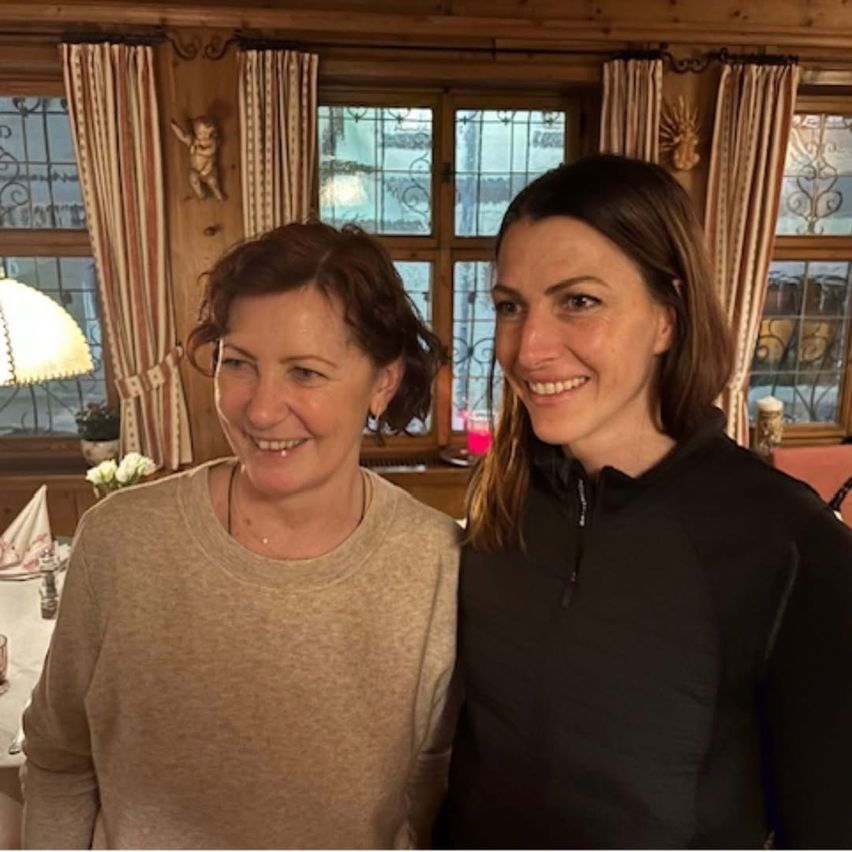 Zwei Frauen lächeln drinnen und stehen neben einem Fenster mit Holzpaneelen und Vorhängen. Die Frau auf der linken Seite trägt ein beiges Oberteil. Die Frau auf der rechten Seite trägt eine schwarze Jacke.