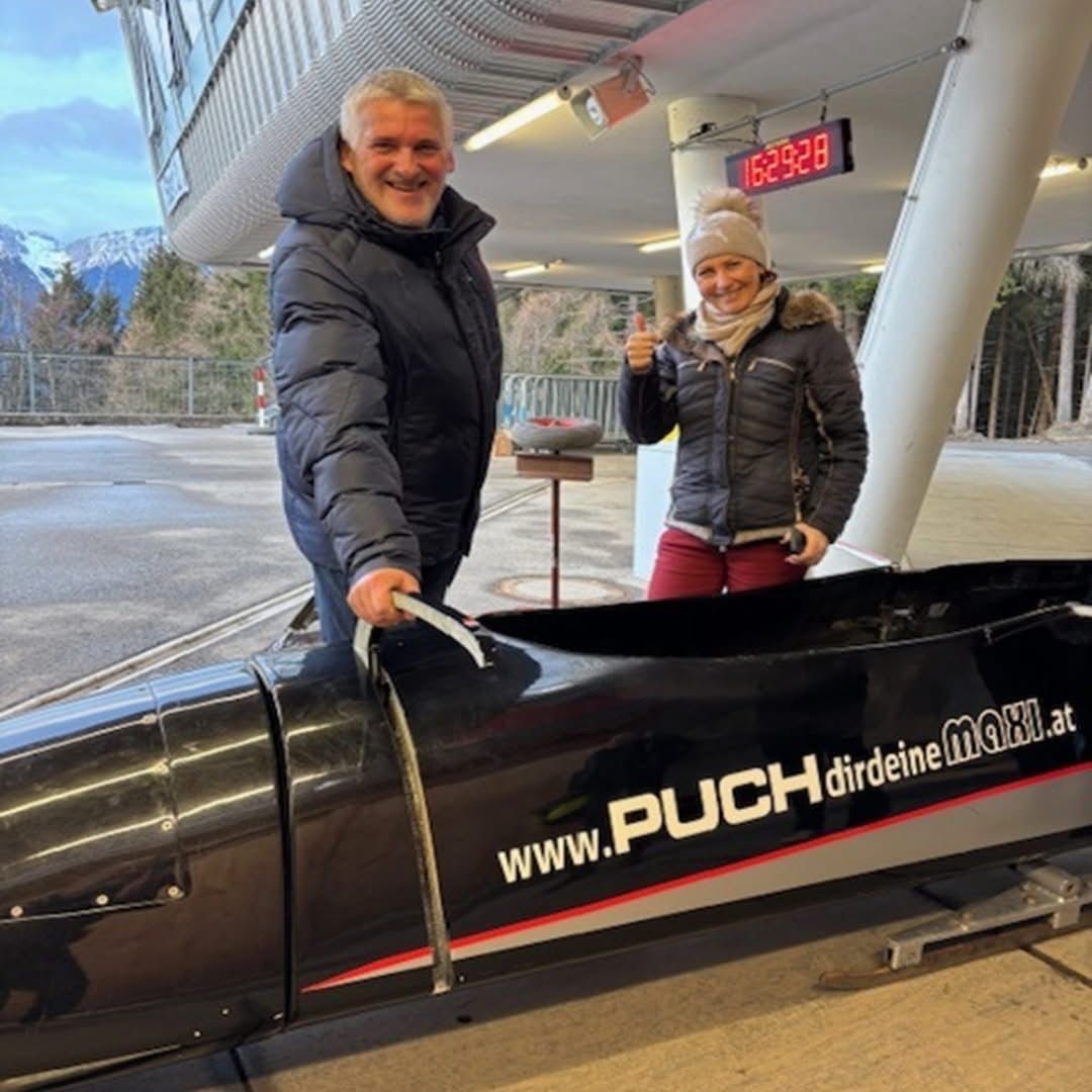 Ein Mann und eine Frau posieren für ein Foto mit einem Bobschlitten an einem verschneiten Ort. Der Mann lächelt, während er den Bobschlitten hält. Die Frau gibt einen Daumen hoch. Der Bobschlitten zeigt eine digitale Uhr und eine Website, www.puchdirdeinemaxi.at.