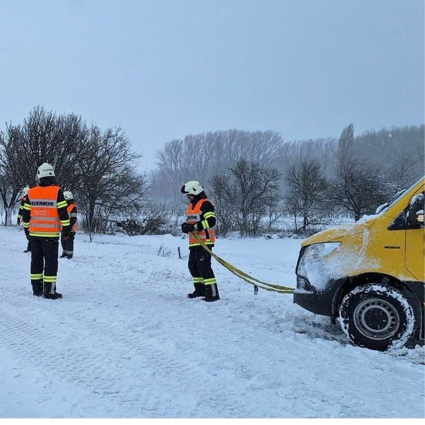 Einsatzkräfte in reflektierenden Westen stehen im Schnee und helfen möglicherweise einem gelben Fahrzeug. Die Bäume sind kahl, und der Himmel ist bewölkt.