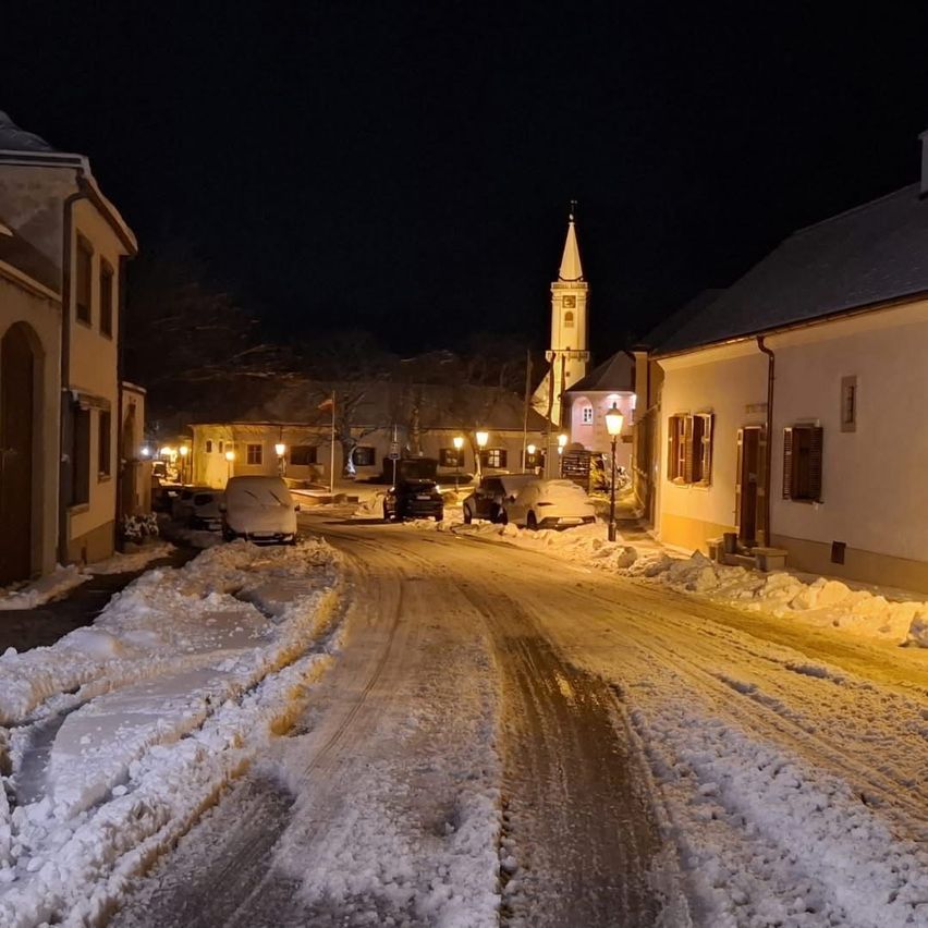 Eine verschneite Straße bei Nacht mit geparkten Autos und Straßenlaternen, die den Weg beleuchten. Eine Kirche mit einem Turm ist in der Ferne sichtbar.