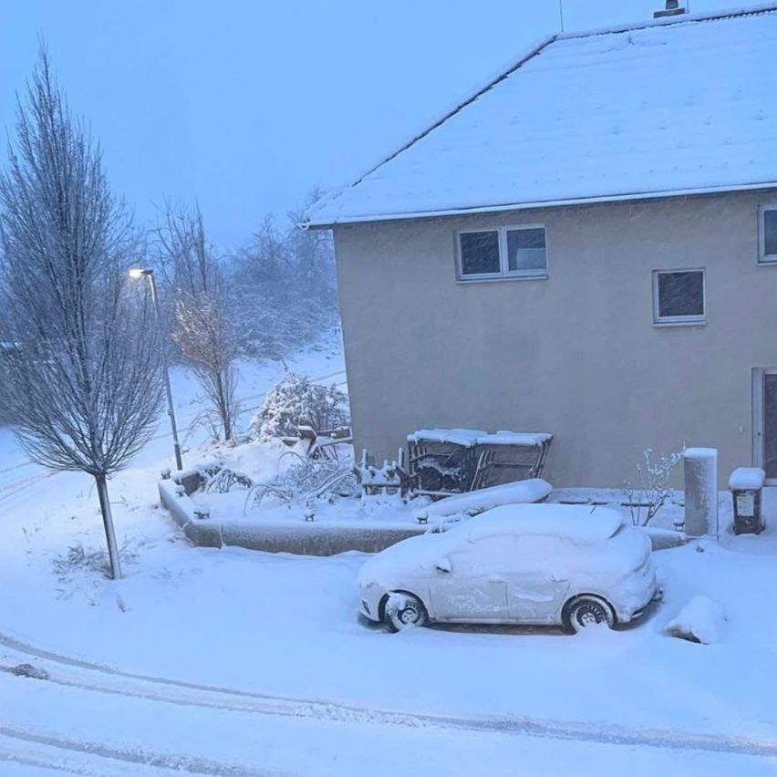 Ein kleines Auto, das in Schnee eingehüllt ist, steht vor einem Haus. Ein schneebedeckter Baum und ein Laternenpfahl sind in der Nähe.