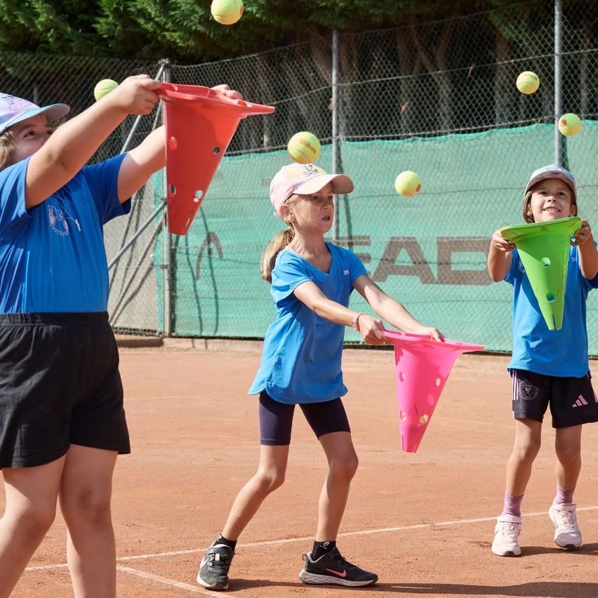 Drei Mädchen spielen Tennis auf einem Platz. Jedes hält einen Kegel und wirft Tennisbälle in die Luft.