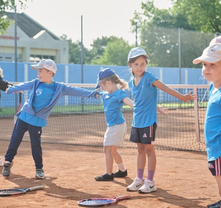 Eine Gruppe junger Kinder in passenden blauen Uniformen steht auf einem Tennisplatz und hält Tennisschläger. Sie üben möglicherweise Tennis.