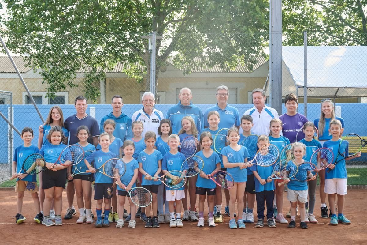 Eine Gruppe junger Kinder und einige Erwachsene in passenden blauen T-Shirts und Shorts posieren für ein Foto auf einem Tennisplatz. Sie halten Tennisschläger in den Händen. Hinter ihnen steht ein Zaun und ein Gebäude.