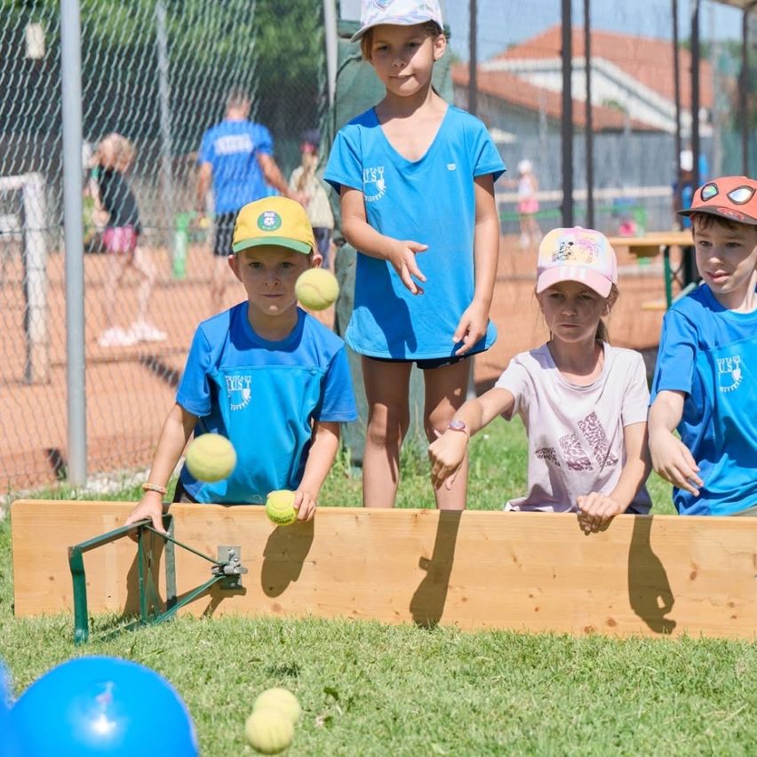 Mehrere Kinder in blauen T-Shirts spielen auf einem Tennisplatz ein Ballspiel, wobei eines einen Ball hält. Hinter ihnen schauen ein Mädchen in einem weißen T-Shirt und ein Junge mit einem Helm auf die Kinder. Im Hintergrund sind andere Leute und ein Gebäude zu sehen.