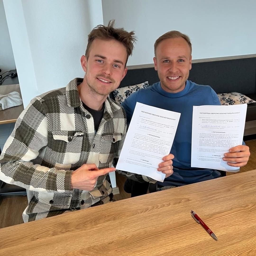 Zwei Männer sitzen an einem Tisch, halten Papiere in der Hand, lächeln und einer zeigt auf das Papier. Ein Stift liegt auf dem Tisch.