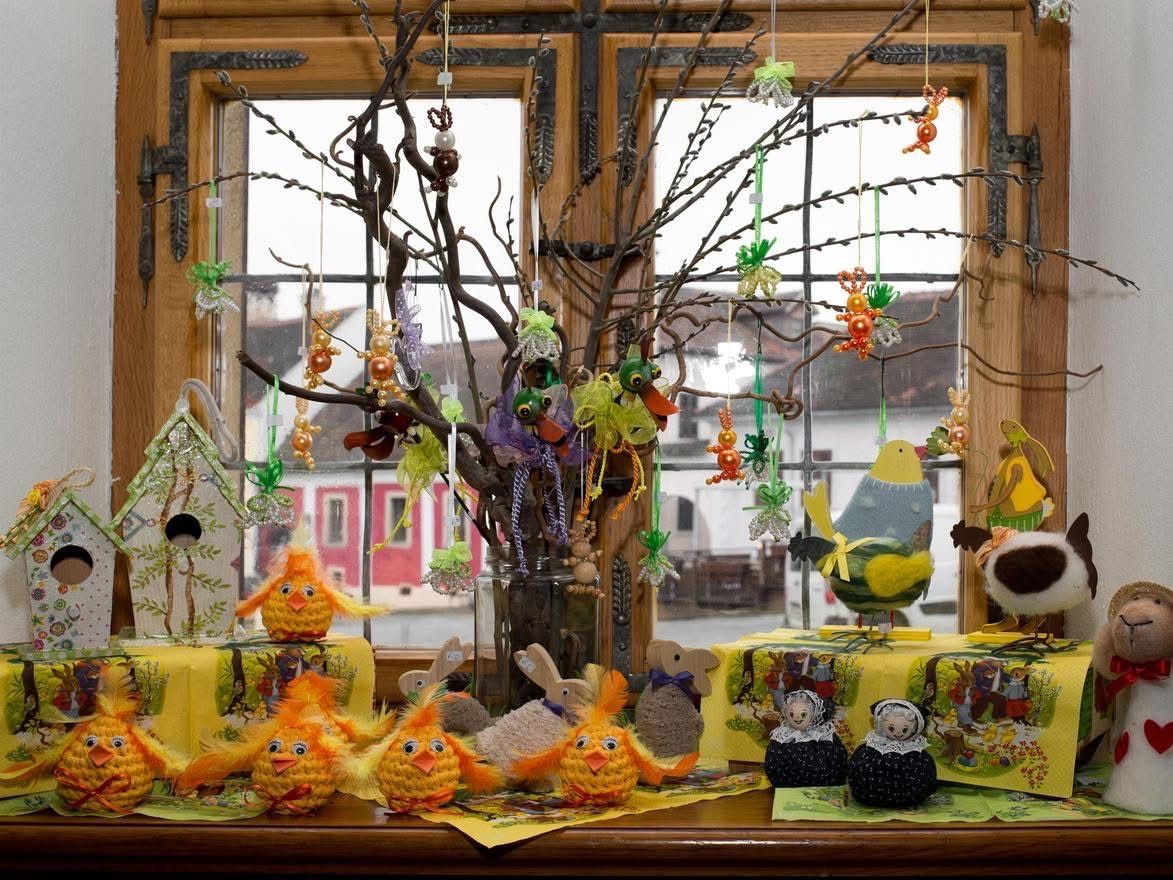 Ein Tisch mit Osterdekoration, einschließlich gehäkelter Küken und Hasen, steht vor einem Fenster mit Blick auf ein rotes Gebäude. Ein Baum mit bunten Ornamenten steht in einer Vase.