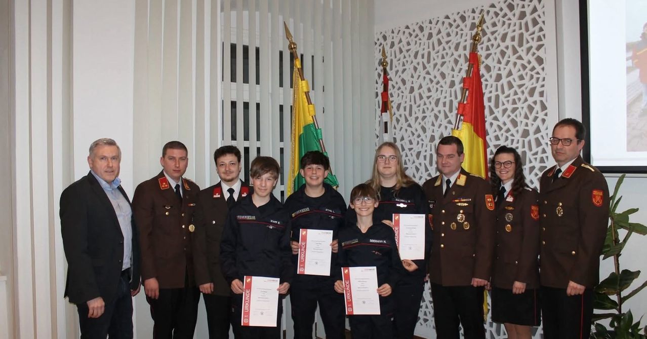 Mehrere Personen in Uniform stehen zusammen und halten Urkunden. Hinter ihnen stehen drei Flaggen, von denen eine grün und gelb ist.