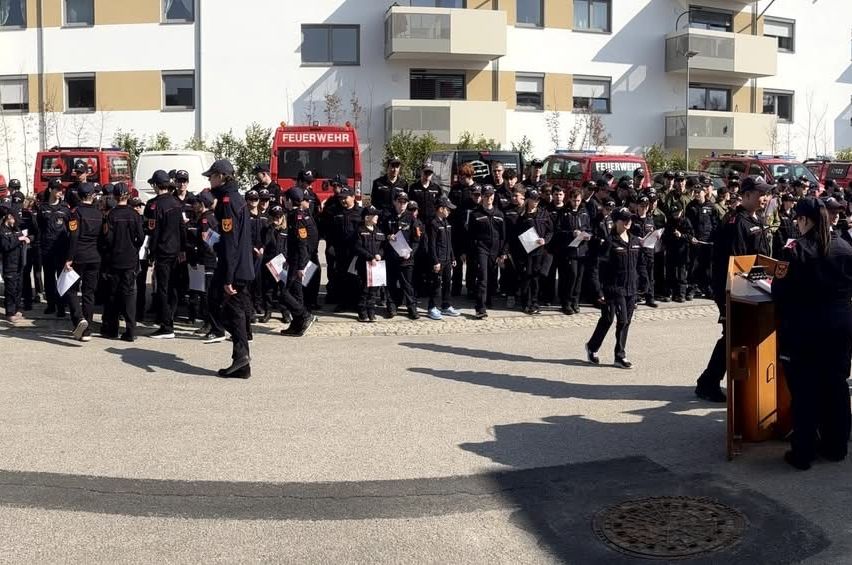 Eine Gruppe von Feuerwehrleuten in Uniform versammelt sich vor einem Gebäude und hält Papiere in den Händen. Im Hintergrund sind Fahrzeuge geparkt.