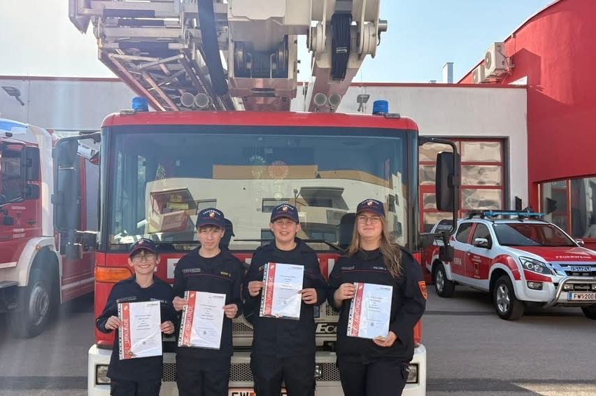 Vier Personen in Feuerwehruniformen posieren vor einem Feuerwehrauto. Sie halten Zertifikate in den Händen, möglicherweise als Abschluss einer Ausbildung. Dahinter befindet sich ein Gebäude und ein rotes Auto.