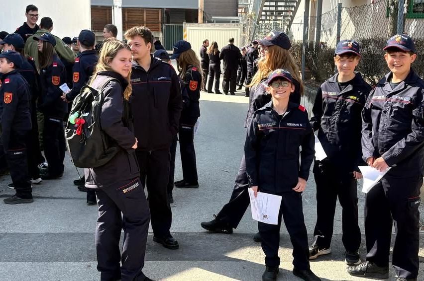 Eine Gruppe von Menschen in Uniformen, wahrscheinlich Feuerwehrleute, stehen in einer Reihe und halten Papiere in der Hand. Sie tragen Mützen, Brillen und Turnschuhe. Sie stehen wahrscheinlich vor einem Gebäude.