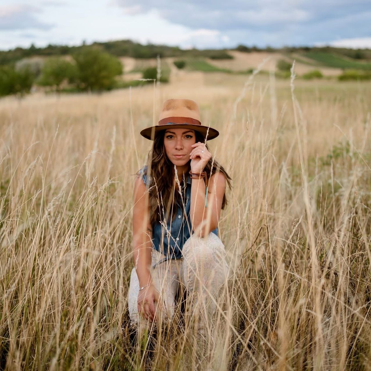 Eine Frau in einem Jeansweste und Hut hockt in einem hohen Grasfeld mit einer entfernten Landschaft im Hintergrund.
