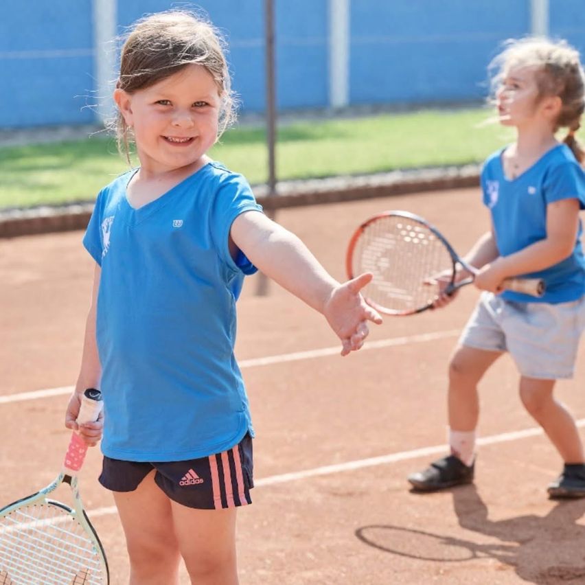 Zwei junge Mädchen in blauen Adidas-Shirts und Shorts spielen Tennis auf einem Tennisplatz. Ein Mädchen lächelt und gibt eine High-Five.