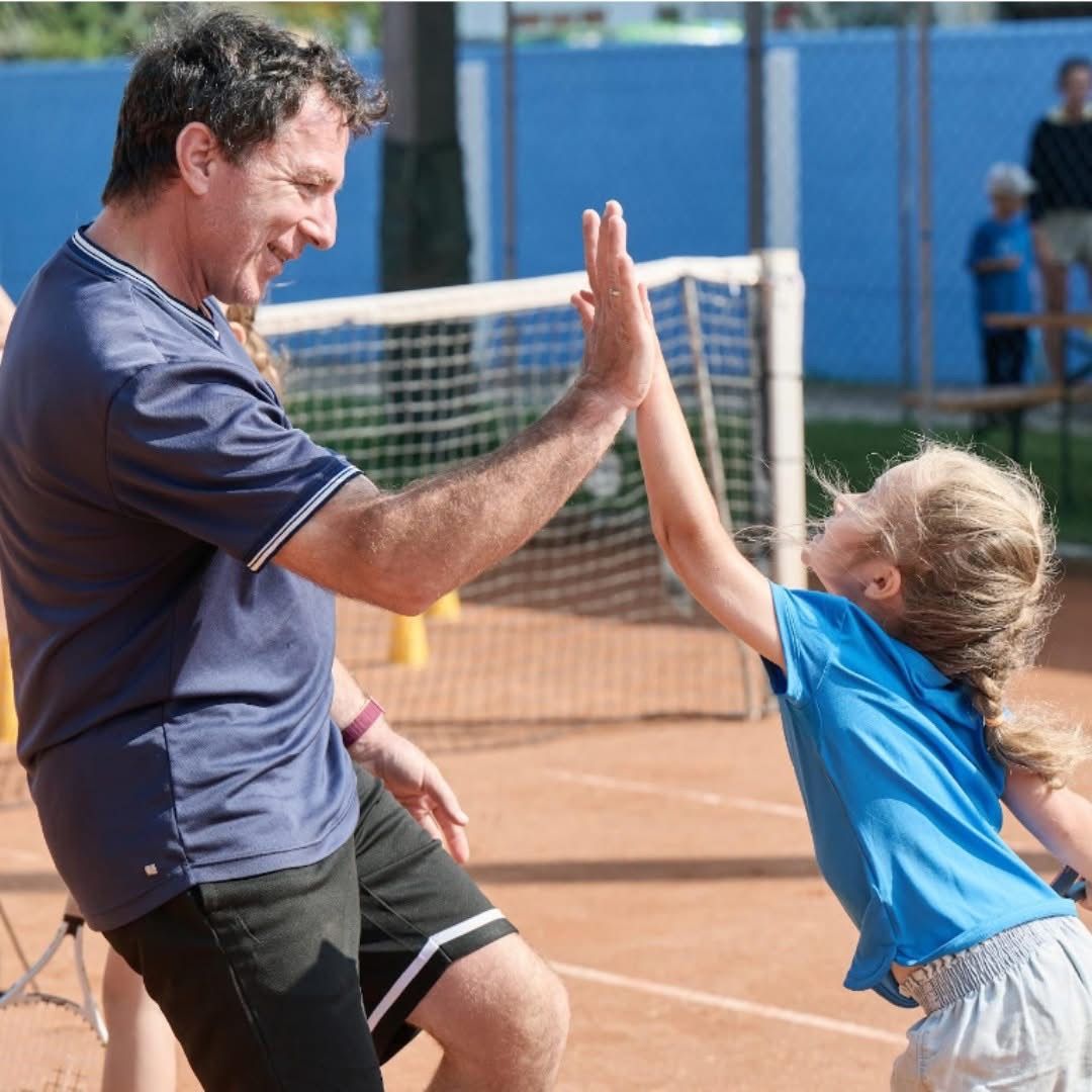 Ein Mann und ein junges Mädchen geben sich auf einem Tennisplatz eine High-Five. Der Mann trägt ein blaues Shirt und schwarze Shorts. Das Mädchen trägt ein blaues Shirt und graue Shorts.