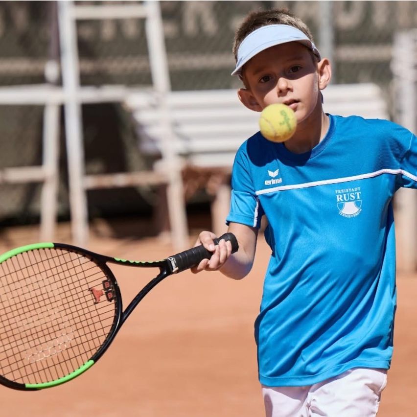 Ein junger Junge in einem blauen Shirt und weißen Shorts spielt Tennis auf einem Sandplatz und hält einen Tennisschläger mit einem gelben Ball in der Luft.