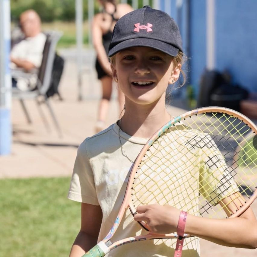 Ein junges Mädchen in einem gelben Shirt und einer schwarzen Kappe mit einem rosa Under Armour Logo lächelt und hält einen Tennisschläger.