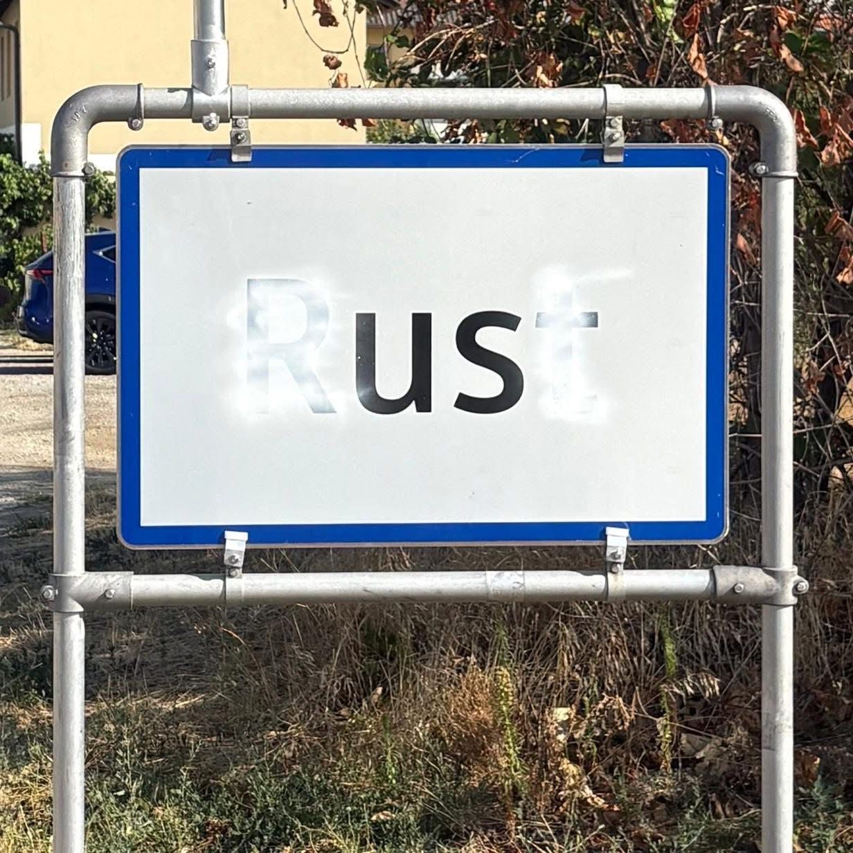 Ein weißes Schild mit blauen Rändern zeigt das Wort 'Rost' in schwarzer Graffiti. Das Schild ist an einem Metallpfosten in einem Grasbereich montiert.