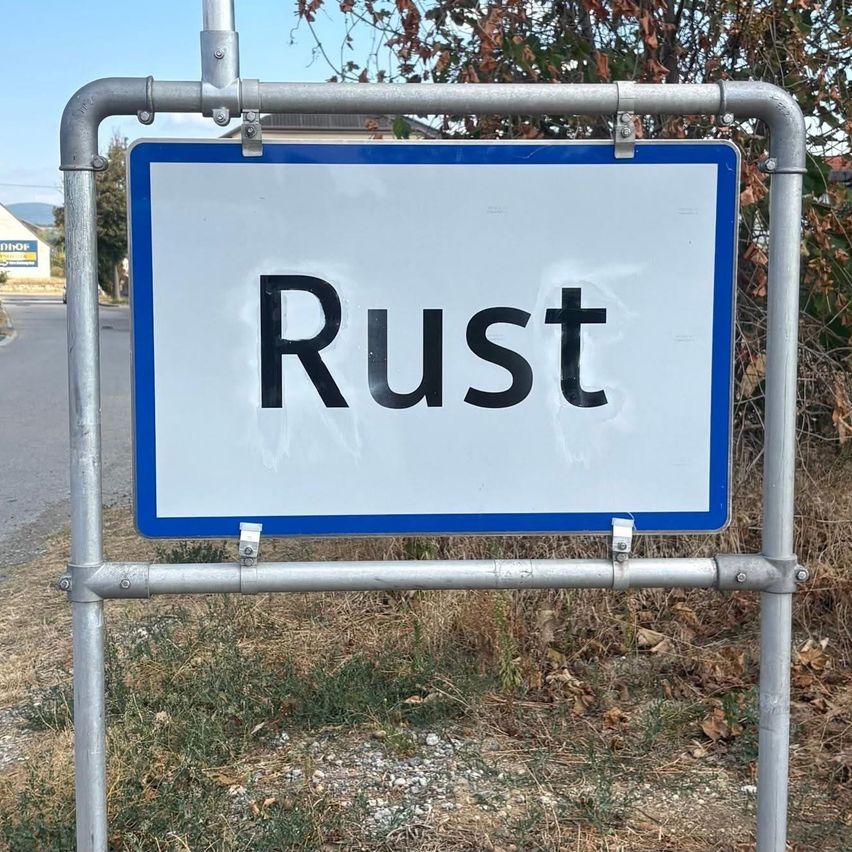 Ein weißes Schild mit dem Wort 'Rost' in schwarzen Buchstaben ist an einem Metallpfosten montiert. Der Hintergrund zeigt einen Grasbereich mit einigen trockenen Blättern und einem Baum.